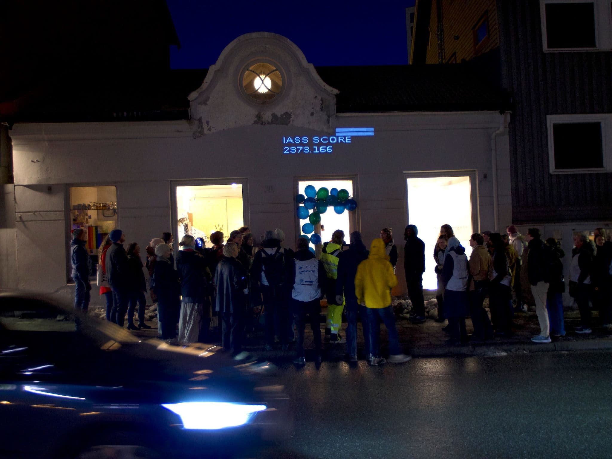 A group of people gathers on a sidewalk at night in front of a building displaying the projected text "IASS SCORE 2373.166" and blue balloons.