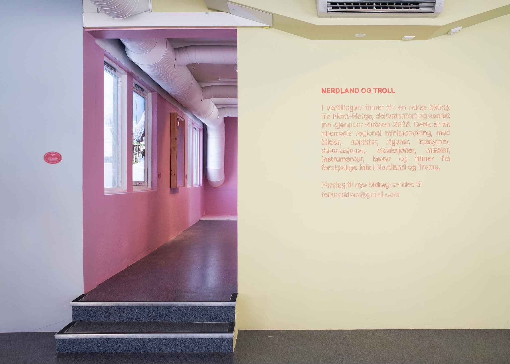 A yellow wall with "NERDLAND OG TROLL" text and exhibition information stands next to a stairway leading into a pink room with visible white ventilation pipes.