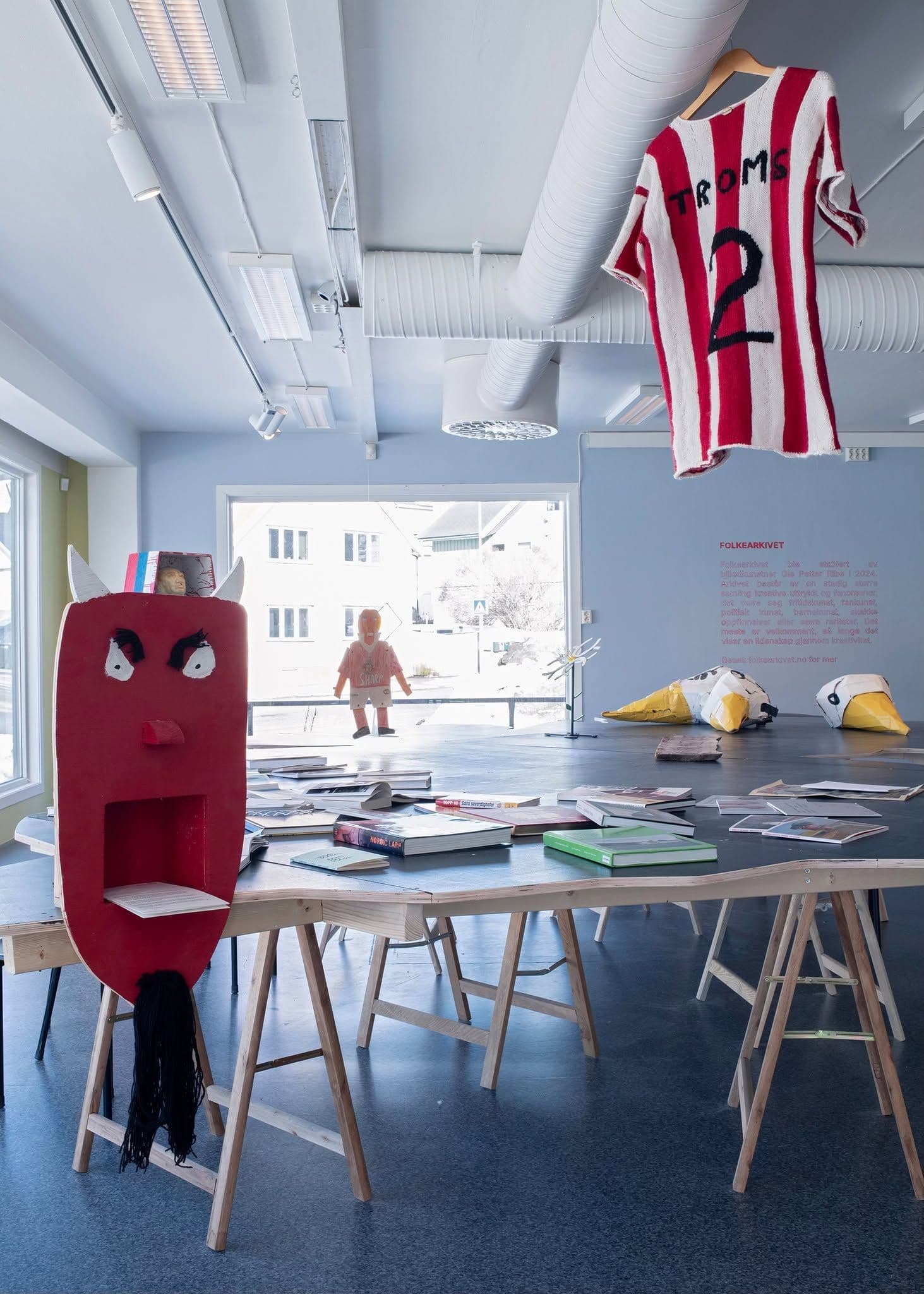 Art installation with a red devil-shaped stand, a hanging sports jersey labeled "TROMS 2", books on a table, and wall text titled "FOLKEARKIVET" in a gallery.