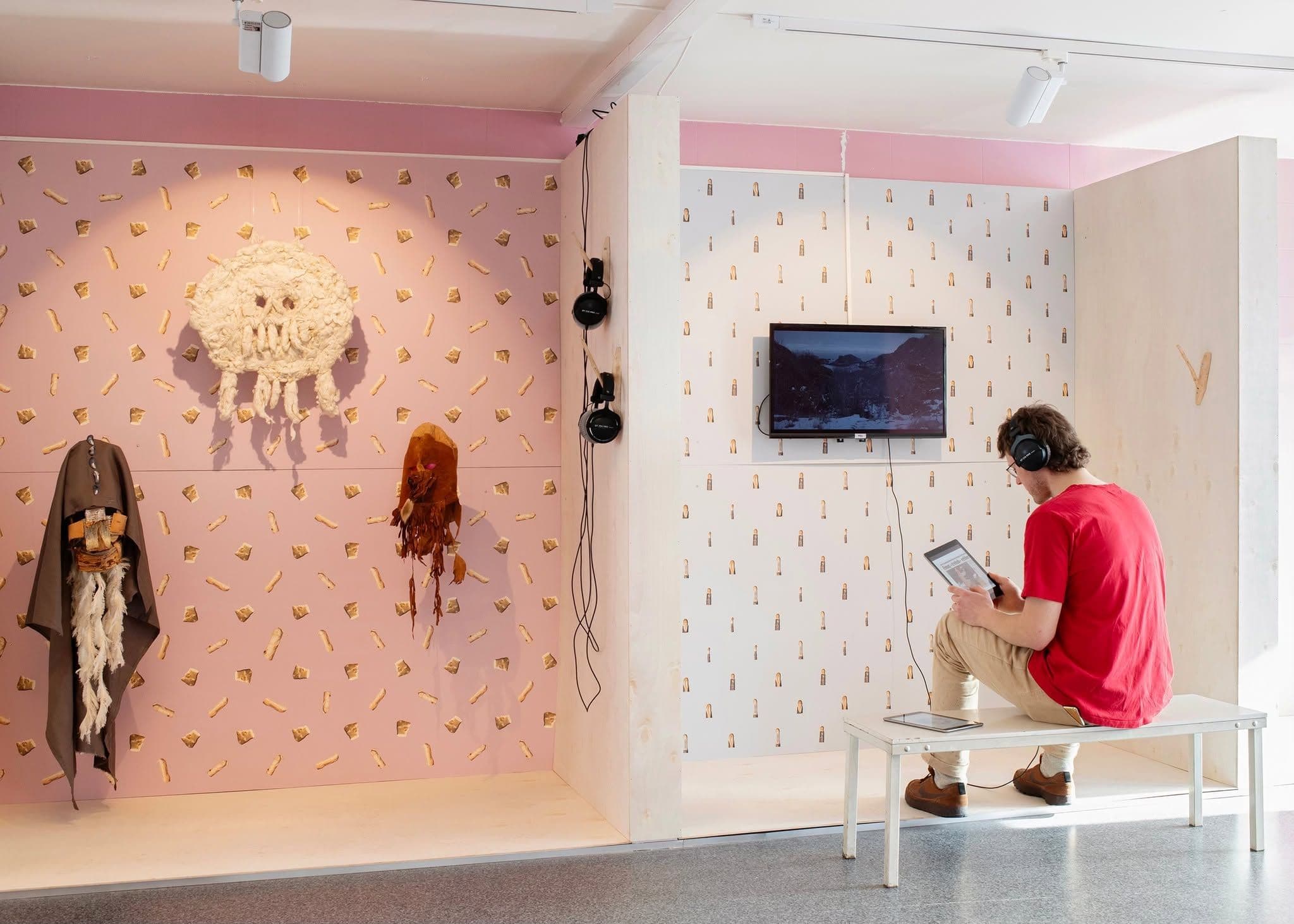 A person in a red shirt sits on a bench, wearing headphones and holding a tablet in an art gallery featuring wall-mounted sculptures and a video screen.