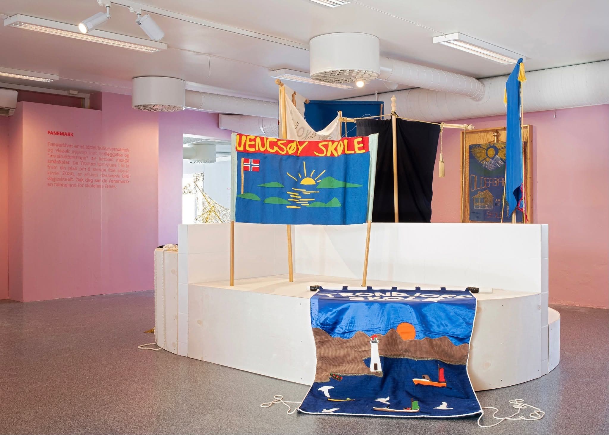 Display of school banners in a gallery space with pink walls; text on a banner reads "VENGSOY SKOLE" above a sunrise, and another reads "TRONDJORD SKOLE".