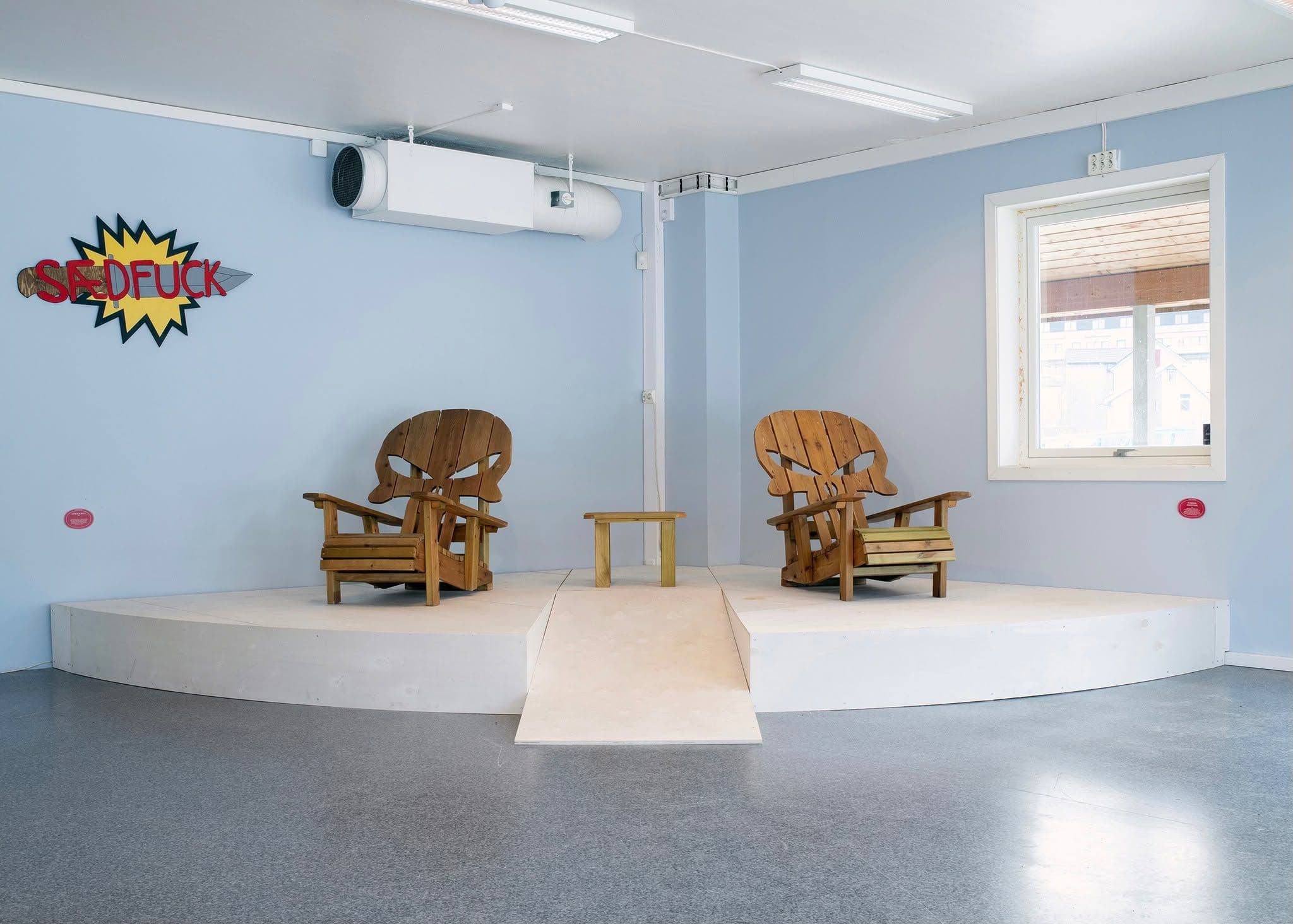 Two wooden Adirondack chairs with skull-shaped backrests sit on a raised white platform in a blue room, with a sign reading "SADFUCK" on the wall.