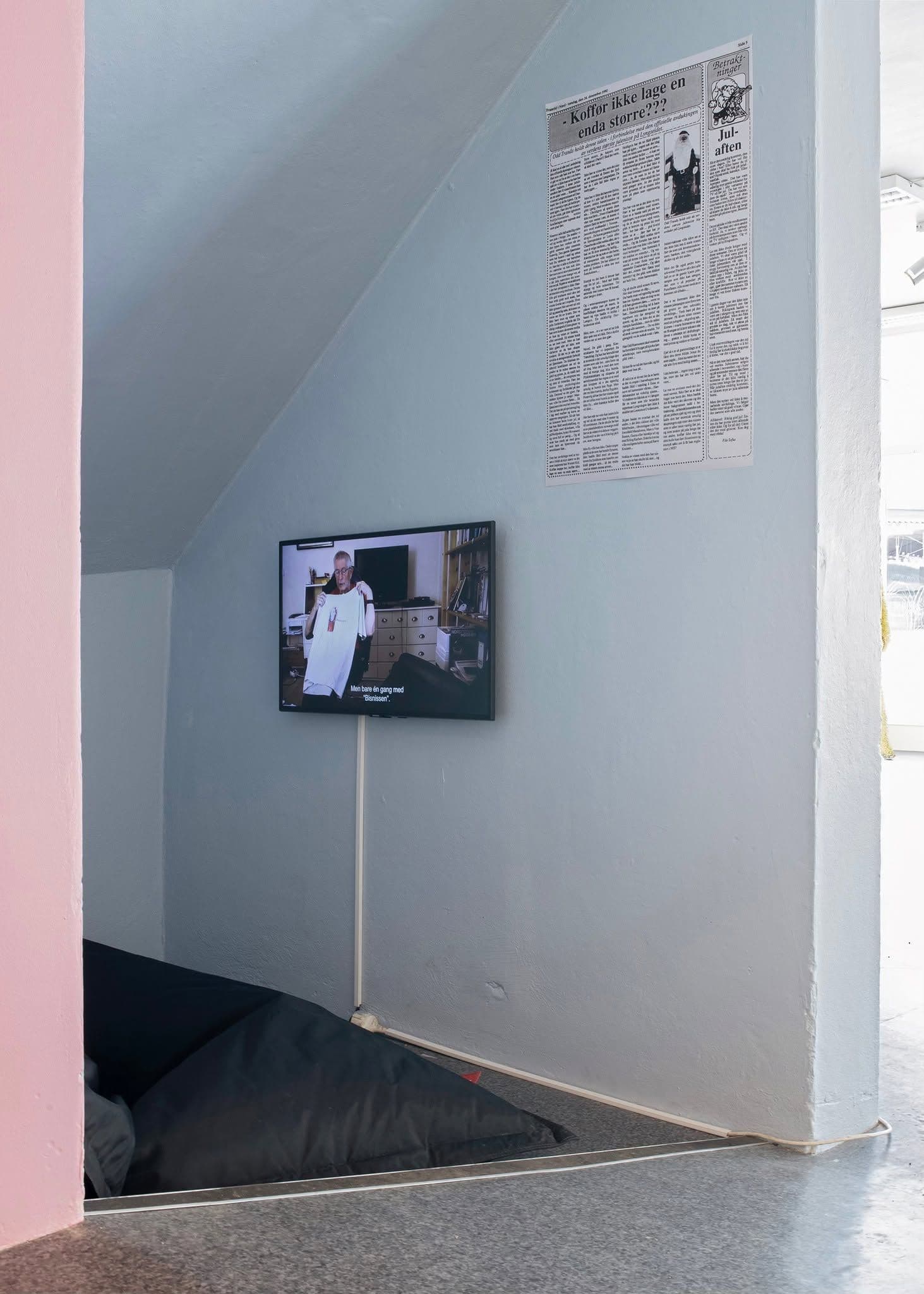 A screen on a gray wall displays a man holding a white shirt, with a large newspaper clipping titled "Koffør ikke lage en enda større???" above it.