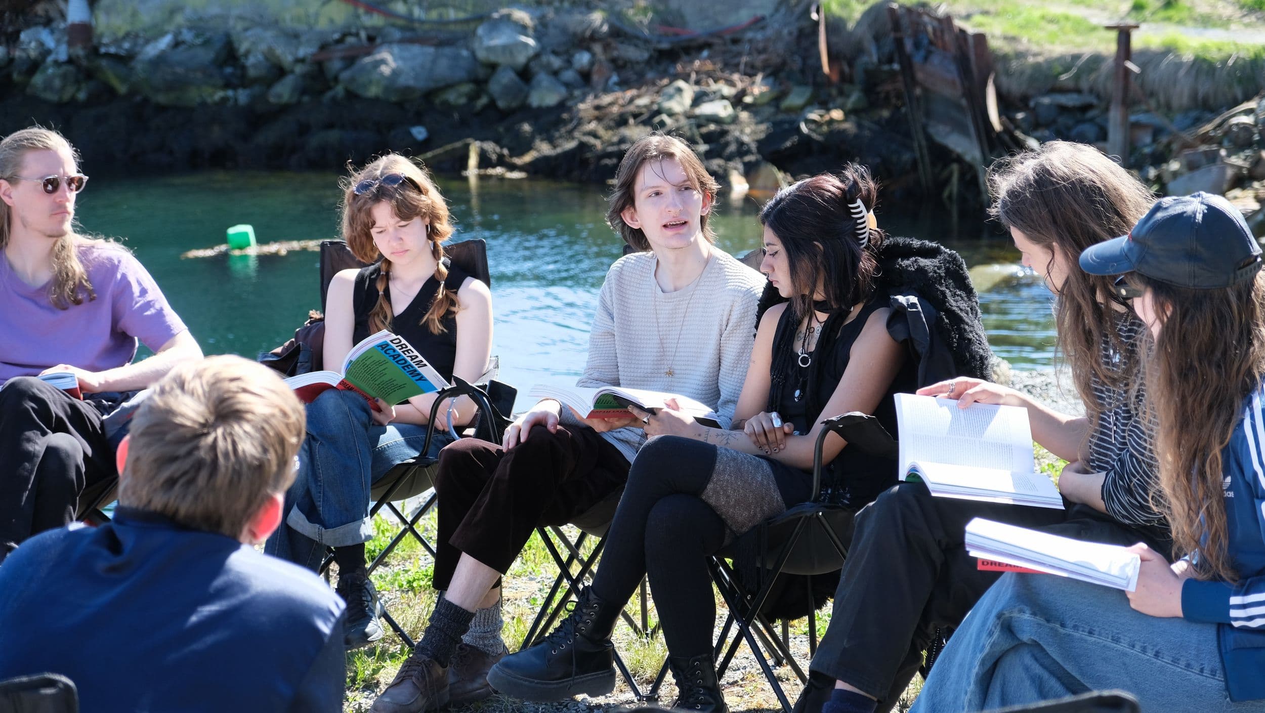 Seven people sit in a circle on folding chairs outdoors by the water, reading books labeled "Dream Academy," while talking to each other.
