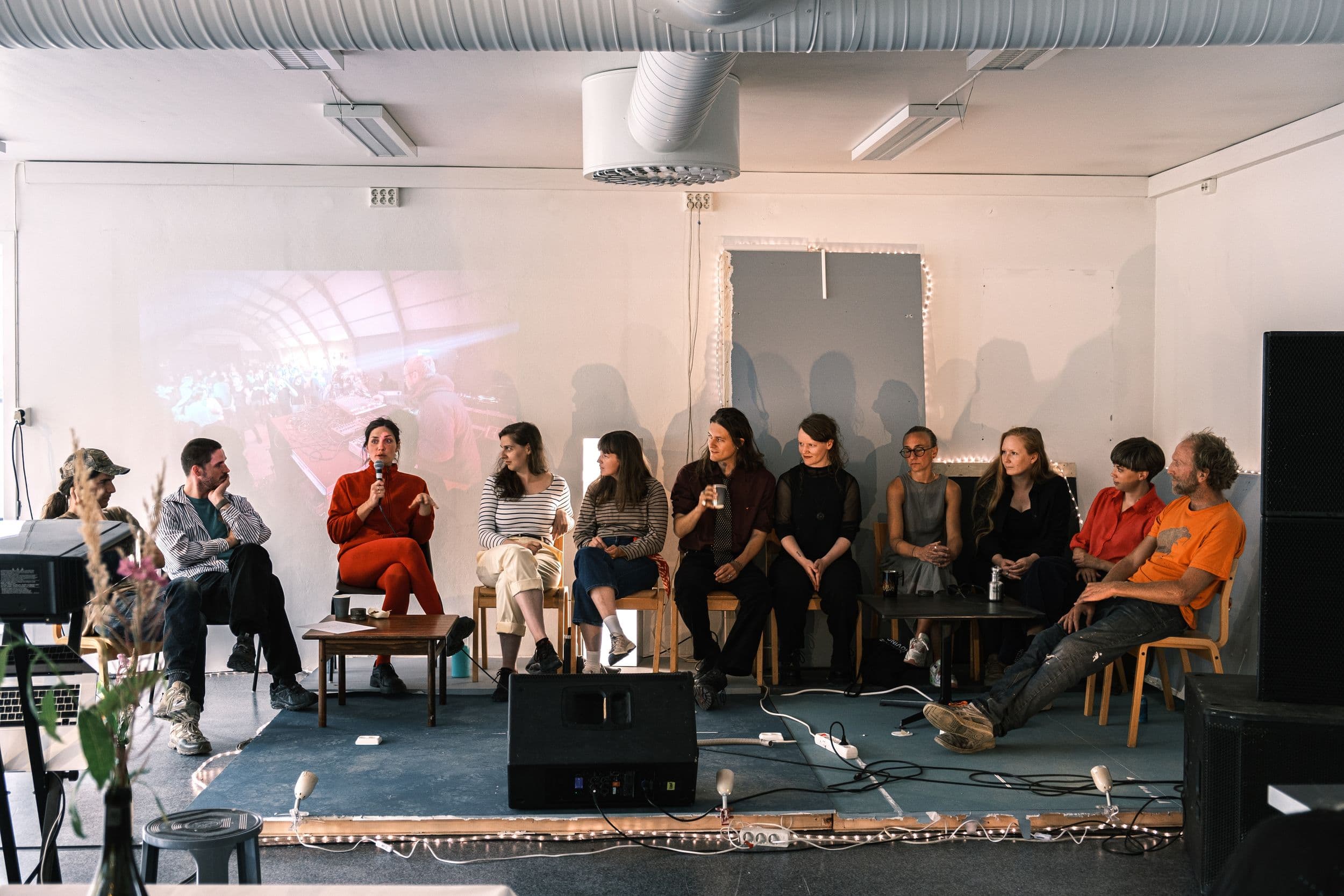 A panel of nine people sits in a row on a raised stage in a brightly lit room, with one person speaking into a microphone while others listen.