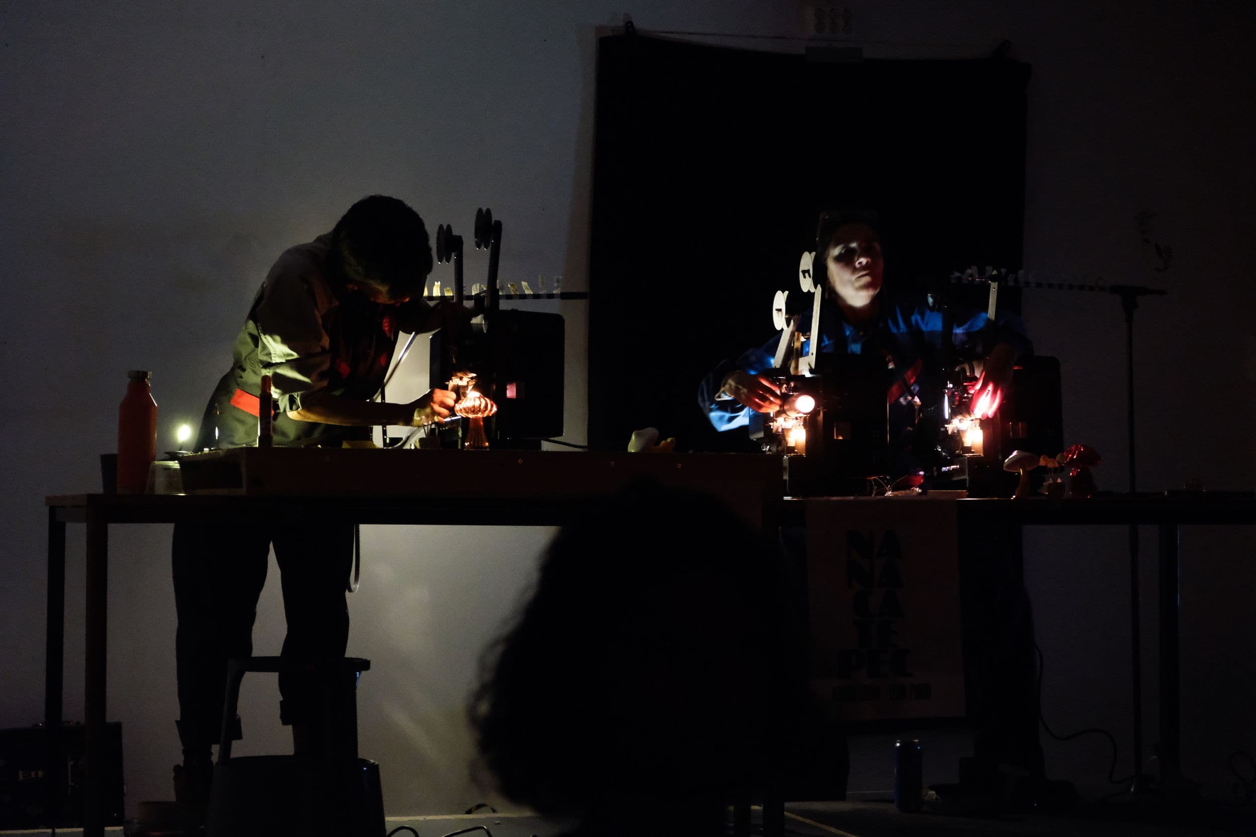Two people operate film projectors on tables in a dimly lit room, with a banner reading "NANACATEPEC" hanging from the right table.