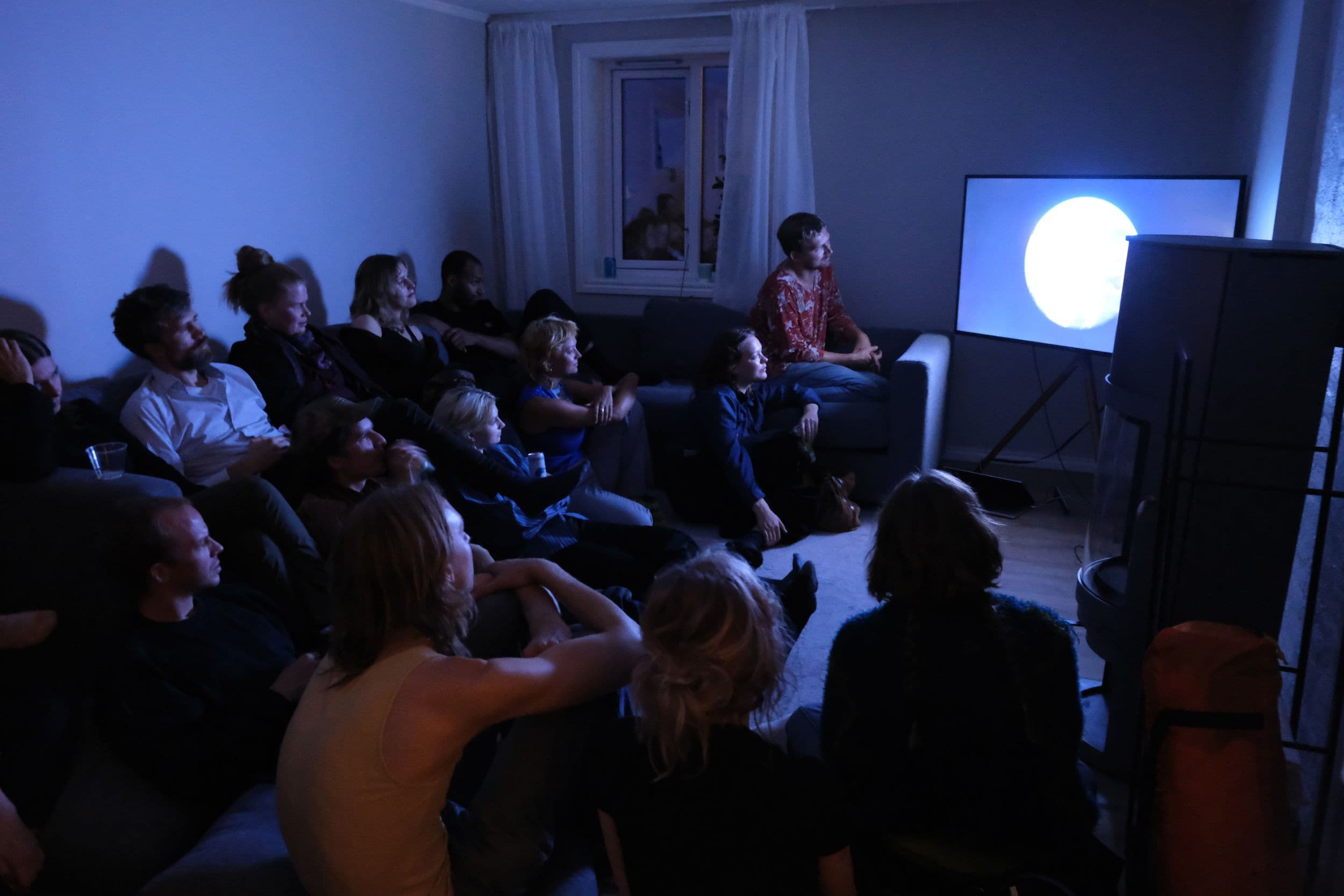 A group of people sits in a dimly lit living room, watching a television screen displaying a bright white circle.