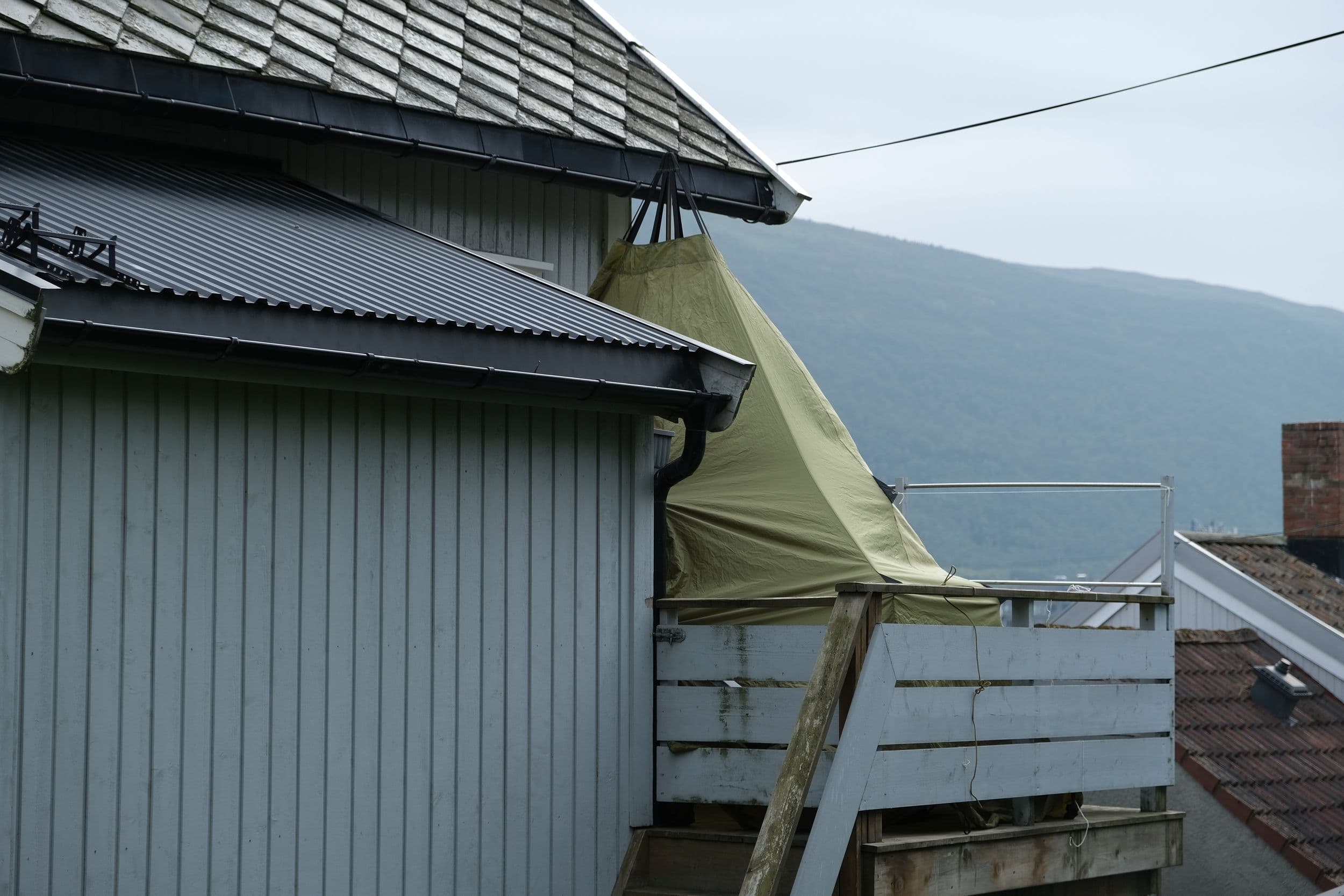 A light green outdoor tent hangs from the eaves of a gray-sided house above a wooden deck railing, with mountains visible in the background.