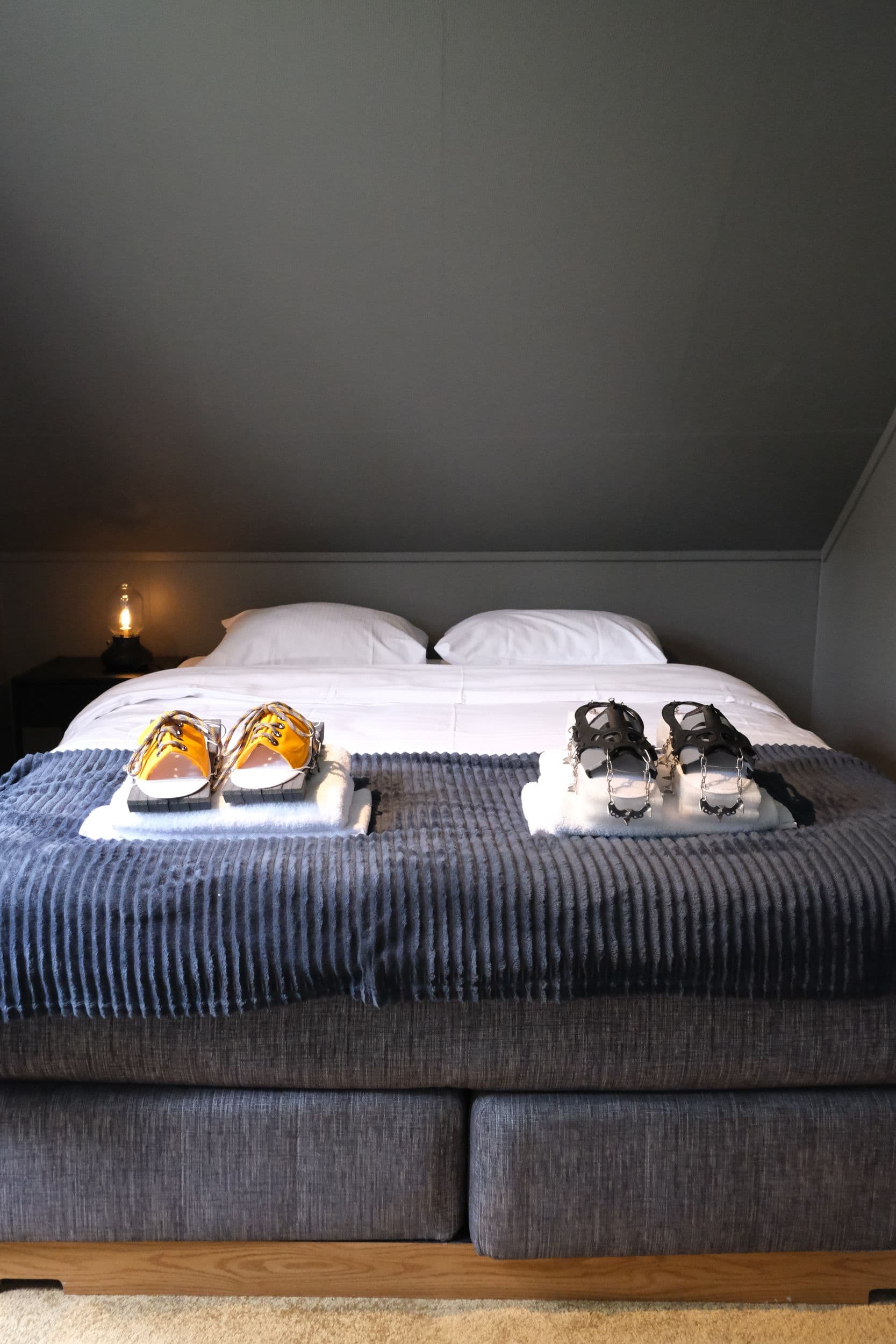 Two pairs of footwear, one yellow and one black with metal spikes, rest on stacks of white towels atop a grey blanket-covered bed under a dimly lit lamp.
