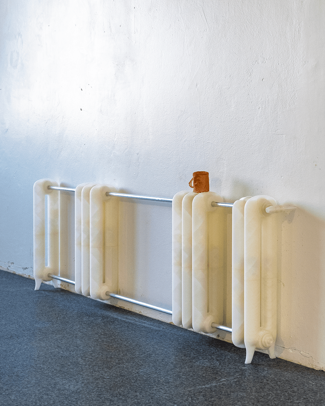 A sculpture of a segmented radiator made of translucent off-white material rests against a white wall on a gray floor, with a small brown cup on top.