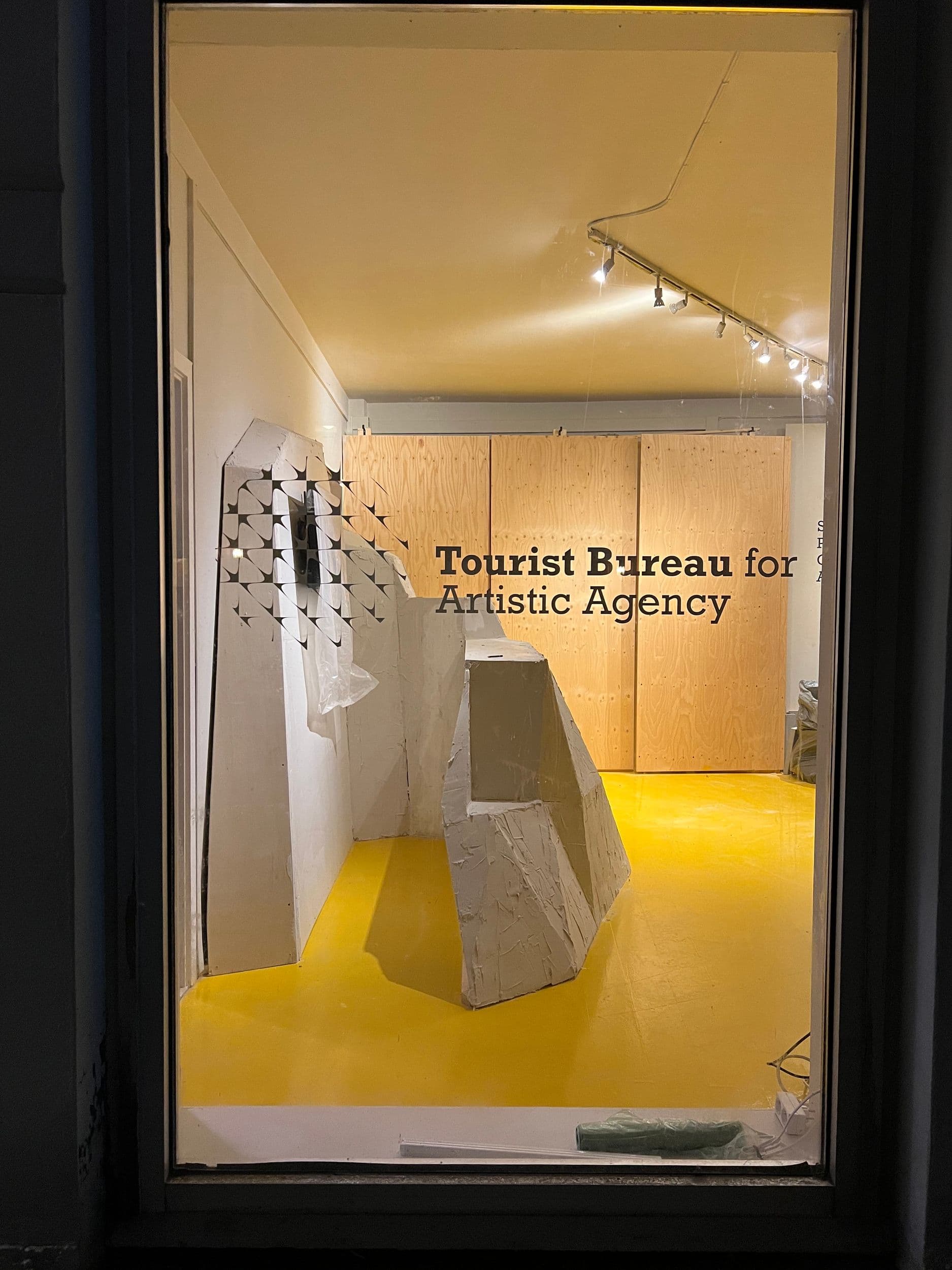 The storefront window displays "Tourist Bureau for Artistic Agency" in text, with geometric white sculptures on a yellow floor against plywood walls.