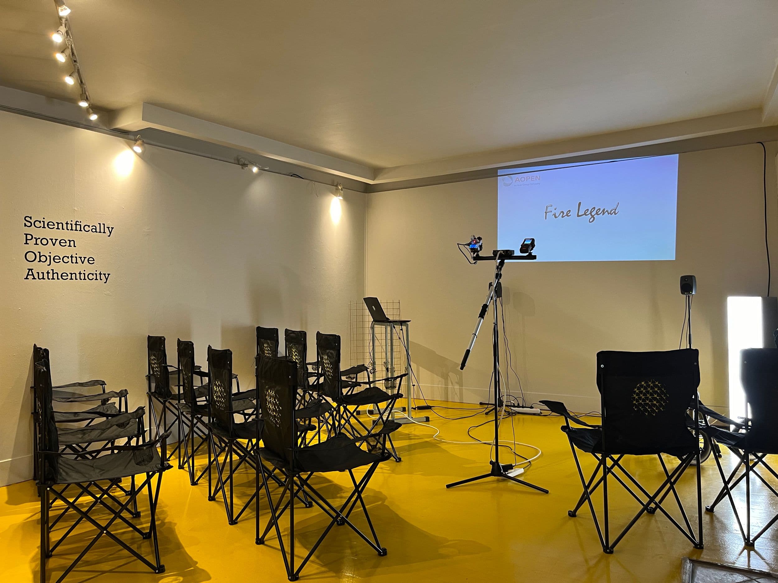 Rows of black folding chairs face a wall projection reading "AOPEN" and "Fire Legend," with text on the left wall reading "Scientifically Proven Objective Authenticity."