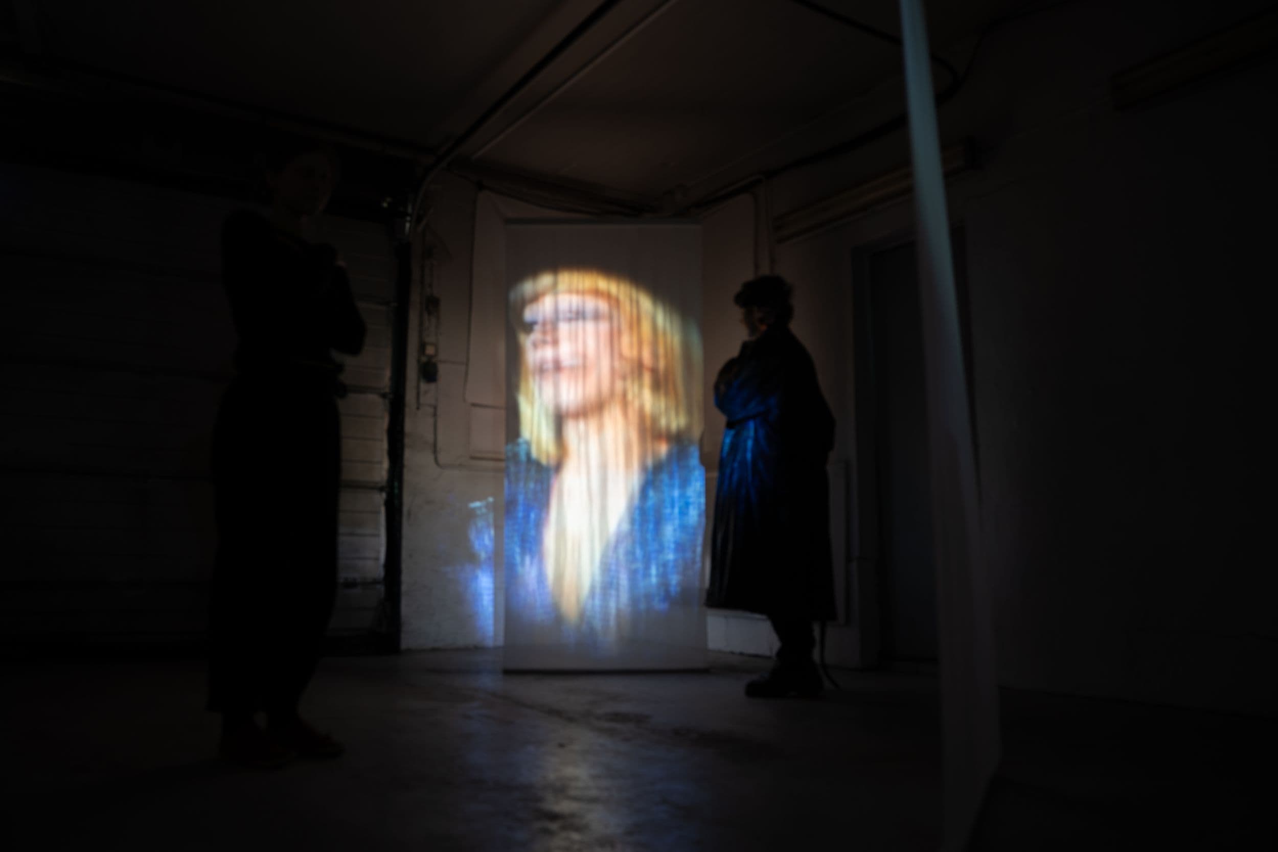 Two silhouettes stand in a dimly lit room, observing a video of a woman projected onto a hanging white fabric panel.
