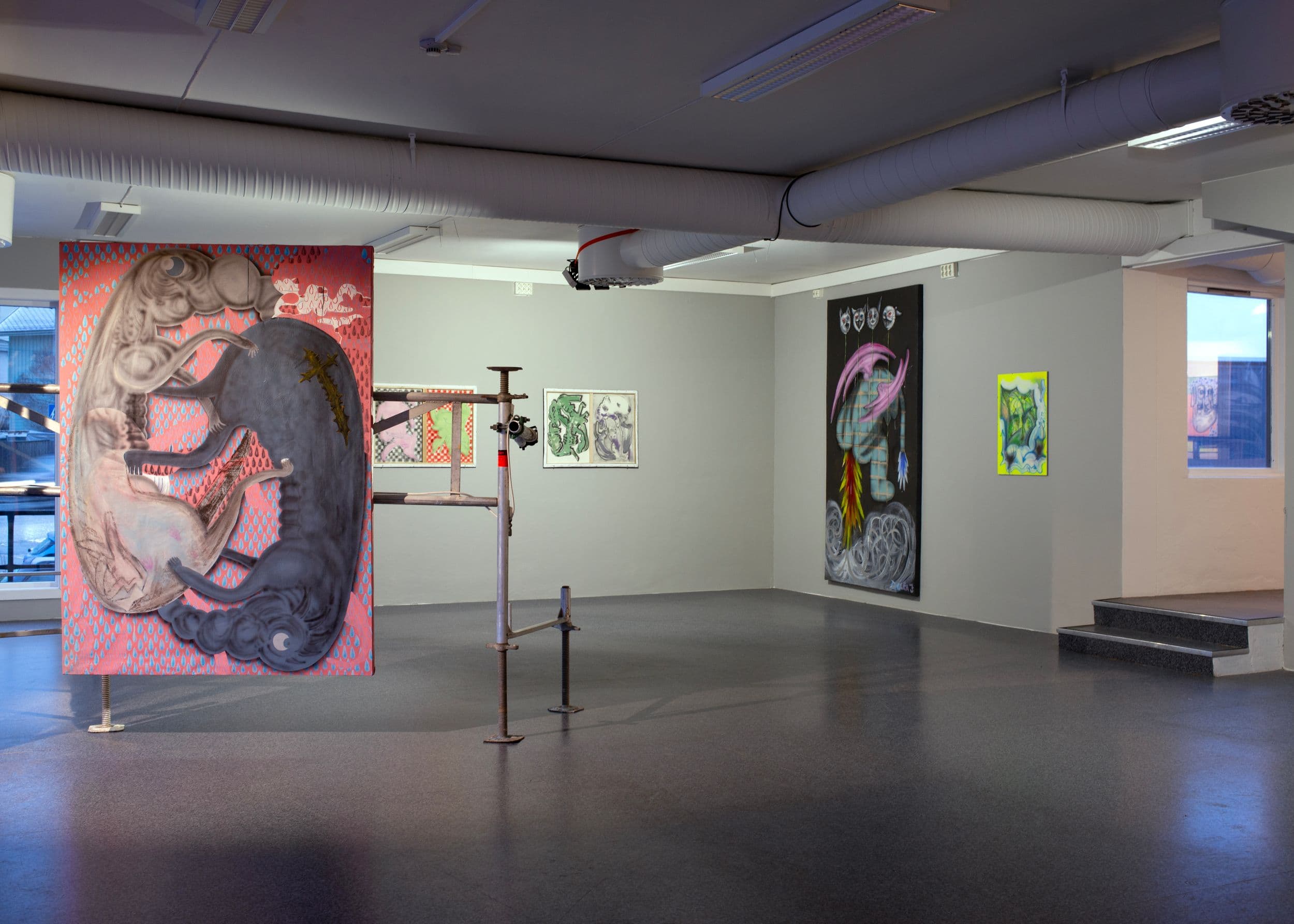 Paintings are displayed on walls and hanging partitions in a gallery space with grey floors and industrial ceiling pipes, including a large work showing creatures.