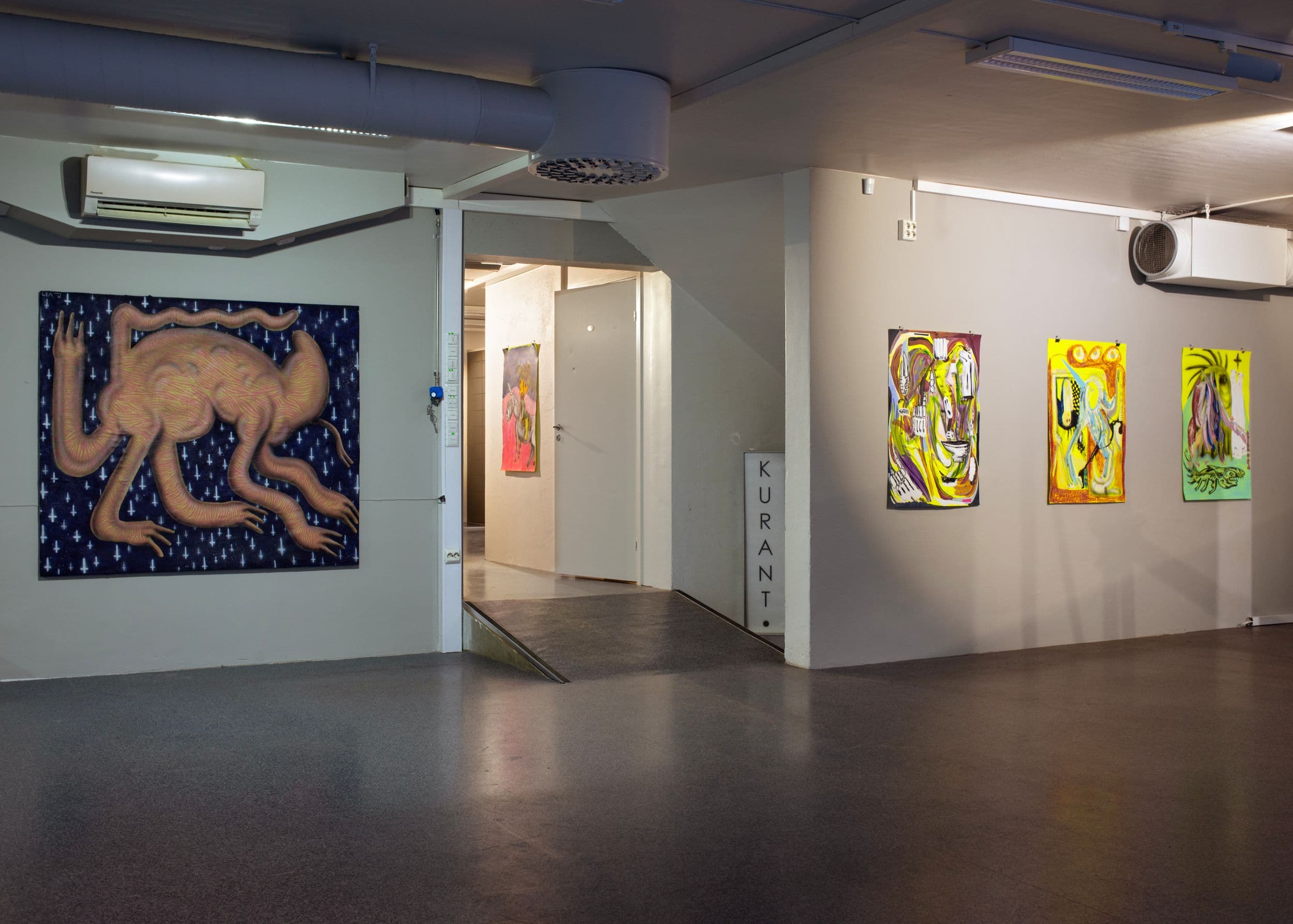 Paintings hang on the walls of an industrial art gallery space, with a vertical sign labeled "KURANT" near a doorway leading to another room.