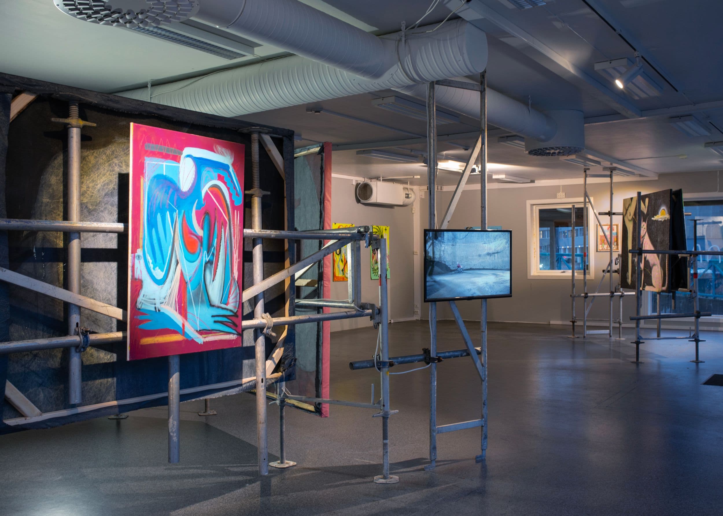 Artworks displayed on scaffolding in a gallery space, featuring a colorful abstract painting on the left and a video screen playing in the center.