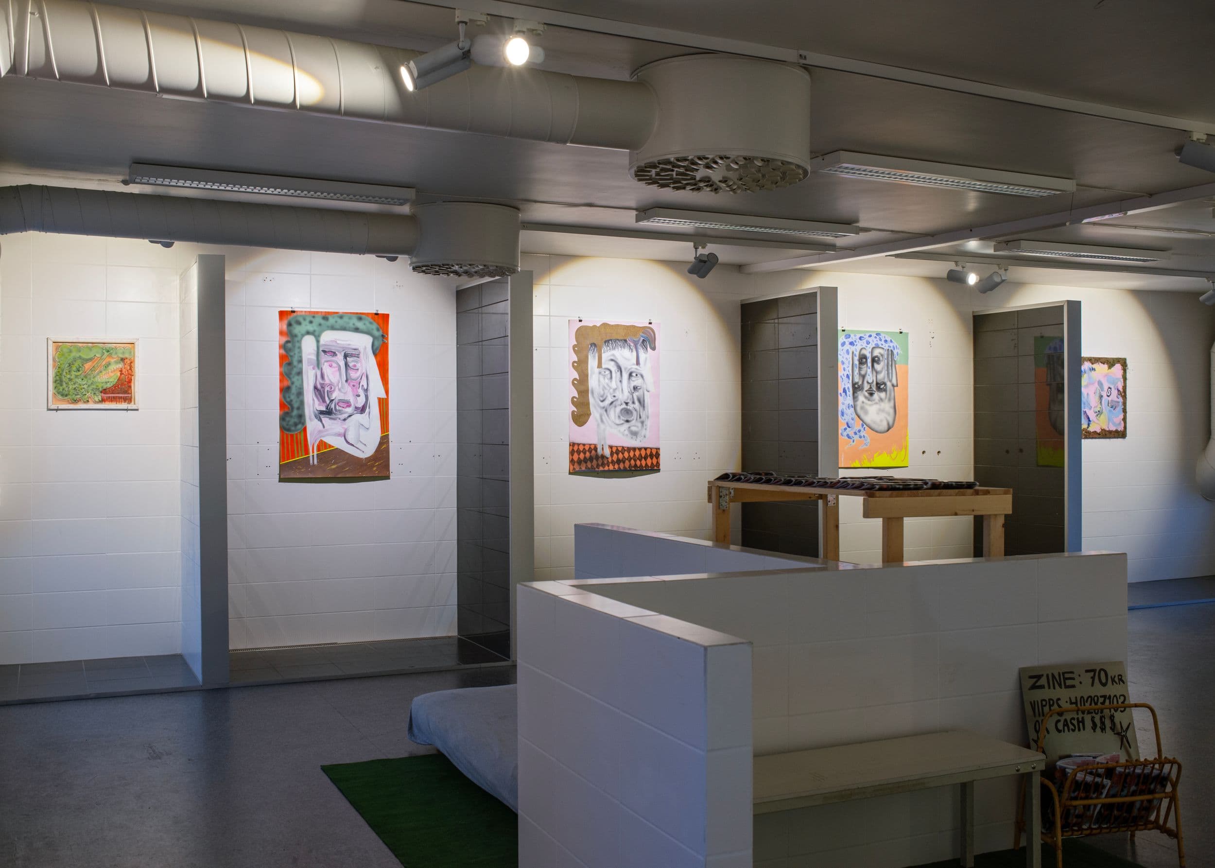 Artworks of stylized faces hang on white-tiled walls in a gallery space with a wooden table, bench, and a sign reading "ZINE: 70 KR VIPPS 40207103 CASH $$".