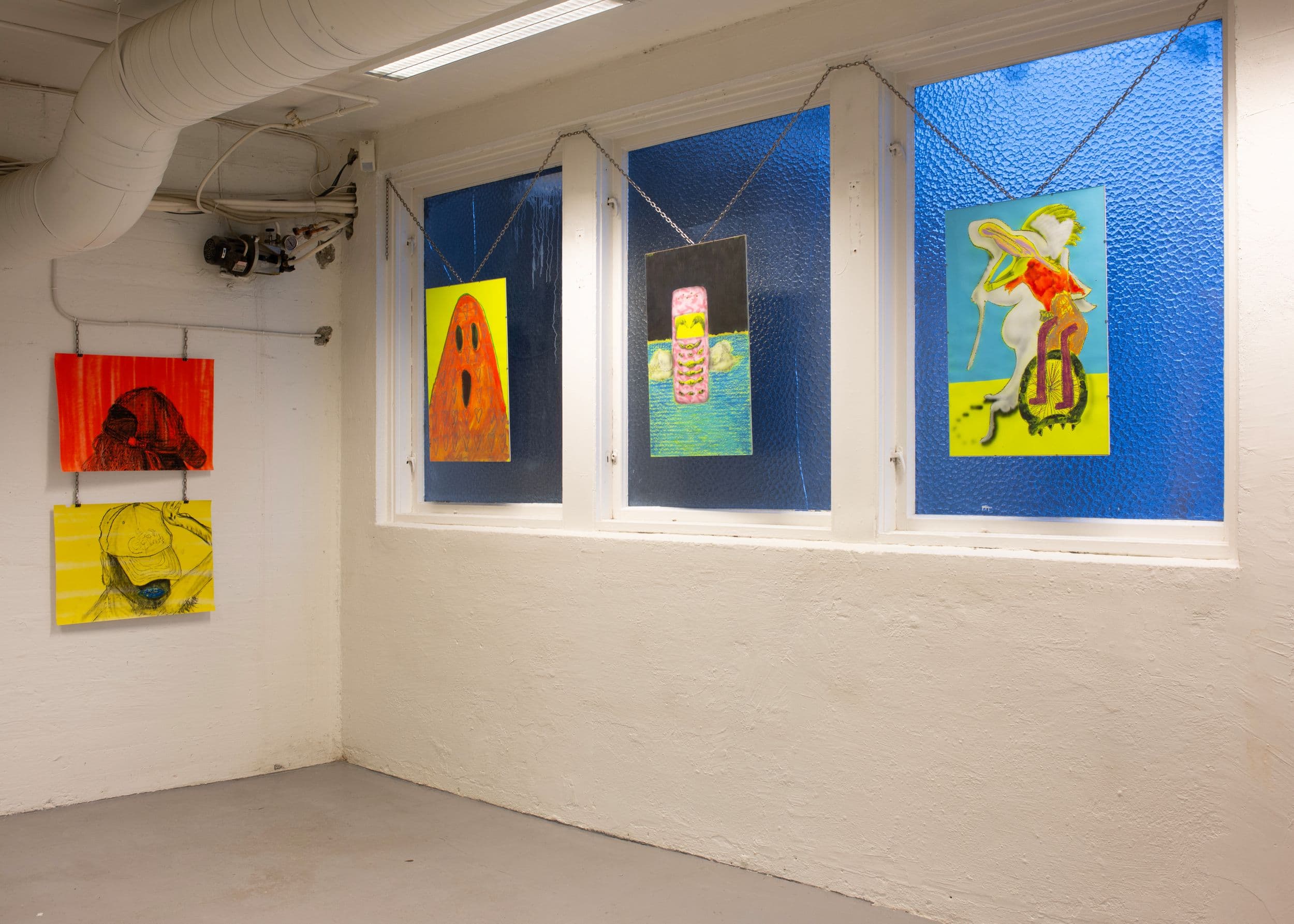 Five colorful, hand-drawn artworks are hung on chains against a white wall and blue-tinted windows in an industrial space.