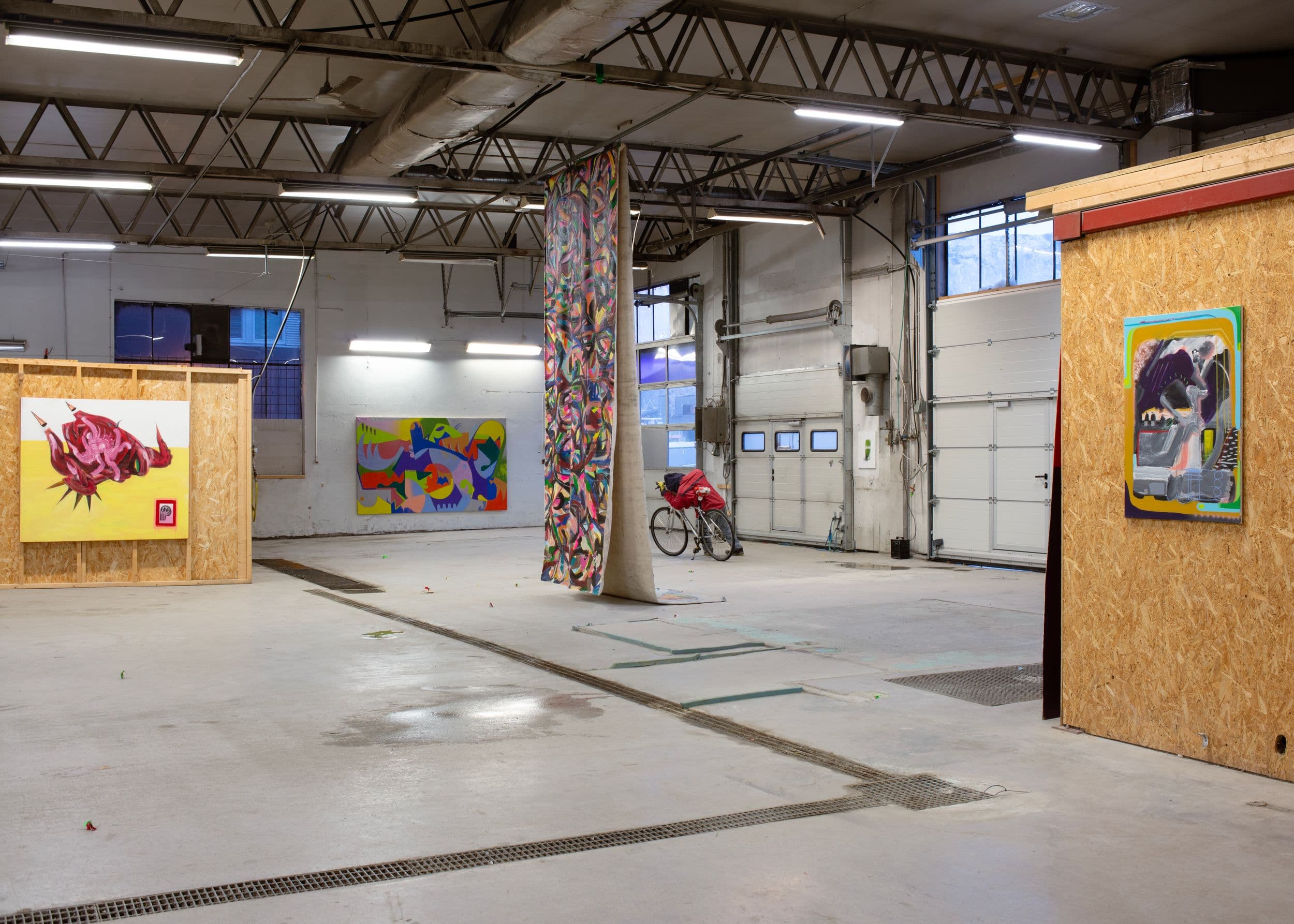 Artworks hang on wooden partitions and white walls inside a spacious industrial warehouse with a bicycle parked near a hanging tapestry.