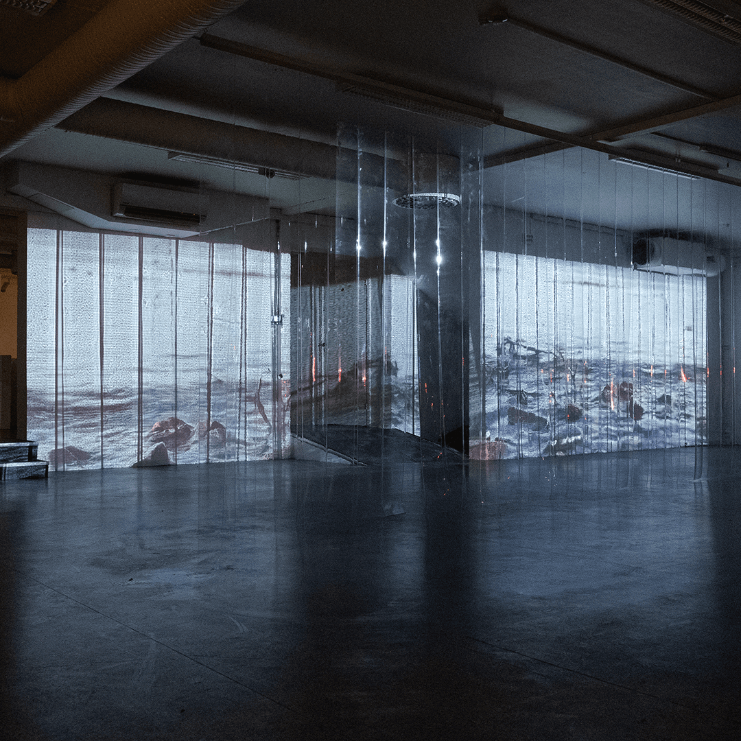 A spacious, dark-floored room features vertical plastic strips hanging from the ceiling, displaying a projected video of a rocky coastline over the interior.