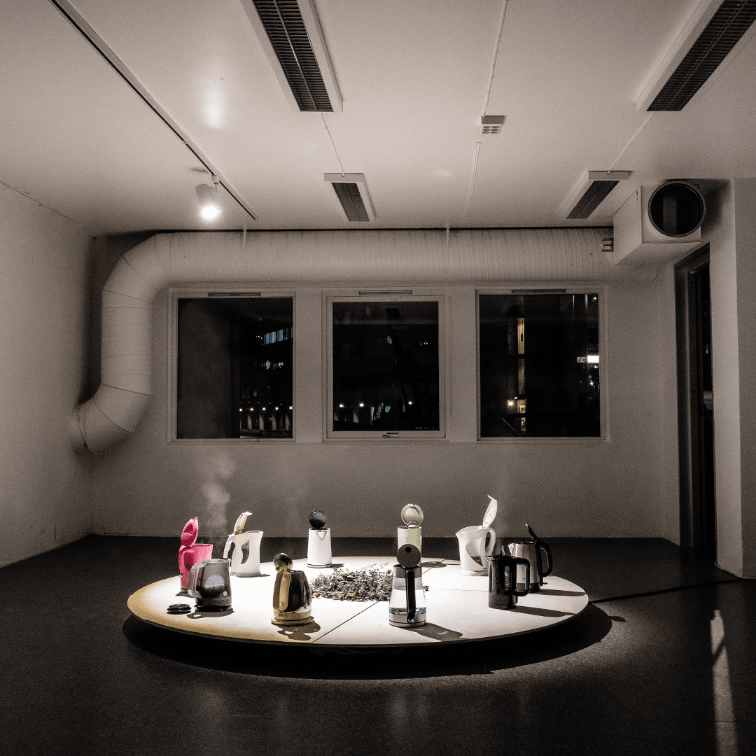 Several electric kettles, some emitting steam, are arranged in a circle on a round platform in a dimly lit, industrial-style room with three windows.