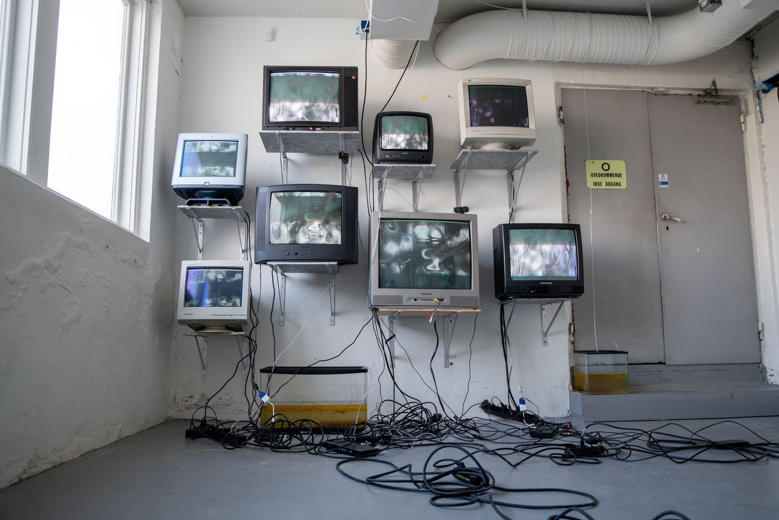 Seven vintage CRT monitors mounted on a white wall display static, with tangled wires leading to two tanks of murky water on the floor and a sign saying "UVEDKOMMEN".
