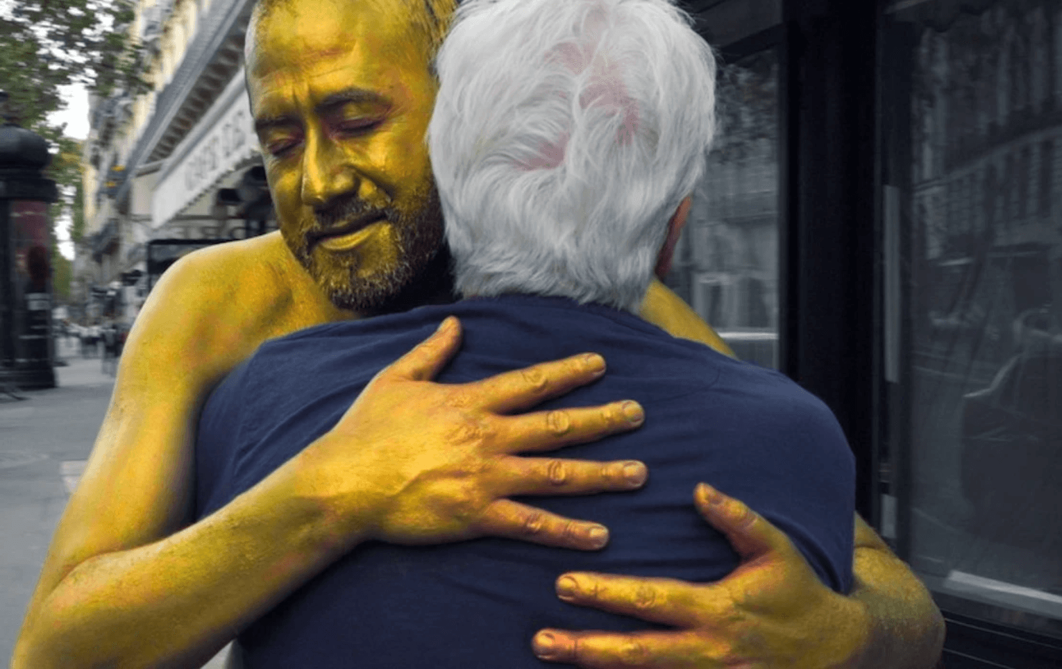 A man covered in gold paint hugs a person with white hair on a city street.